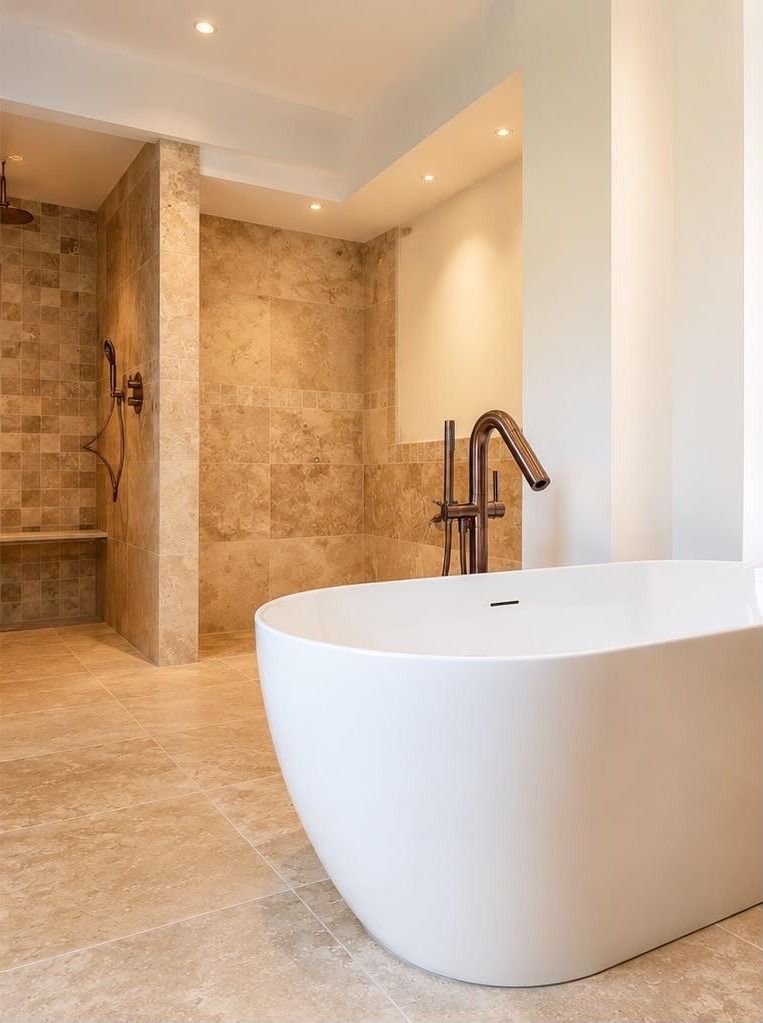 Baignoire îlot blanche avec robinetterie sur colonne finition cuivre, devant une douche à l’italienne carrelée en travertin, réalisée par ENP