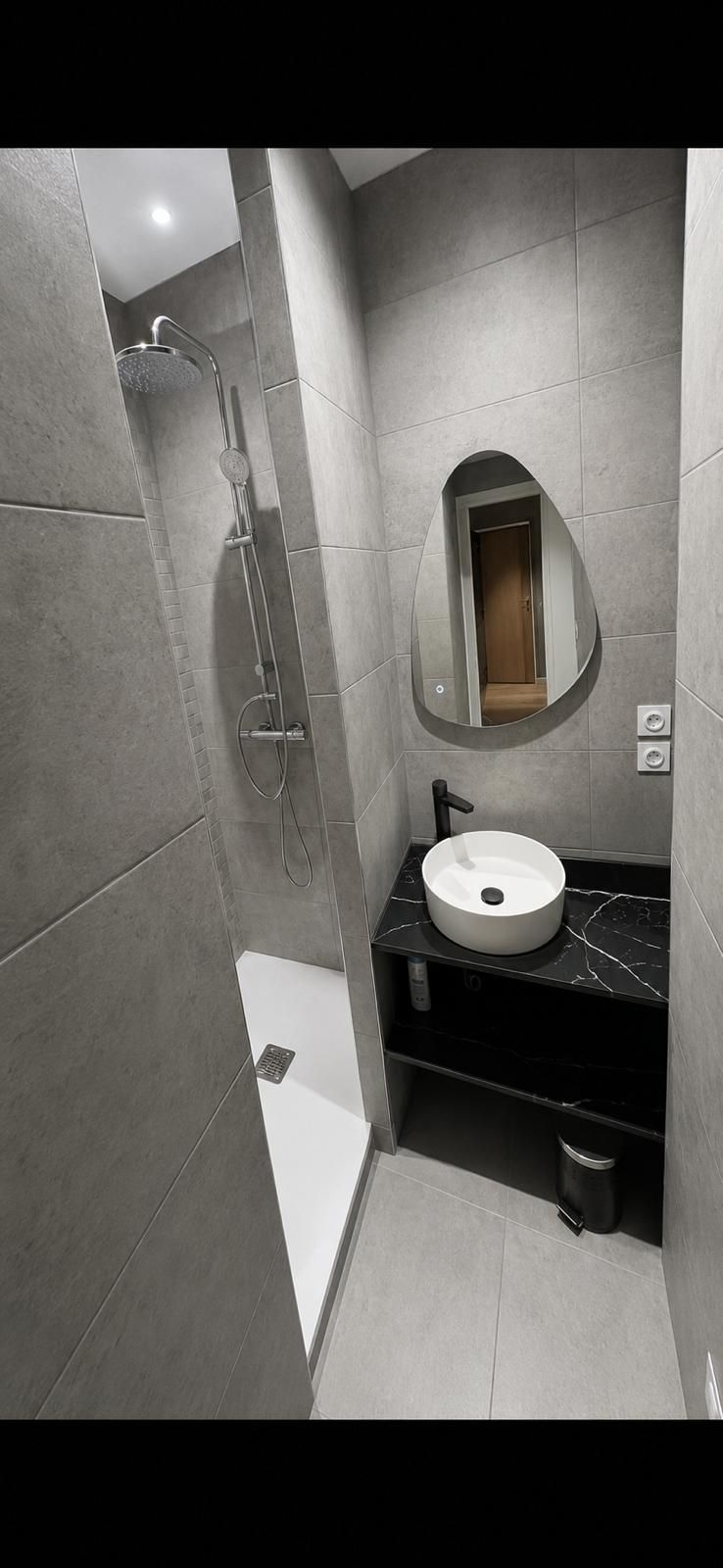 Salle d’eau étroite avec douche italienne carrelée en gris, vasque ronde posée sur plan en marbre noir